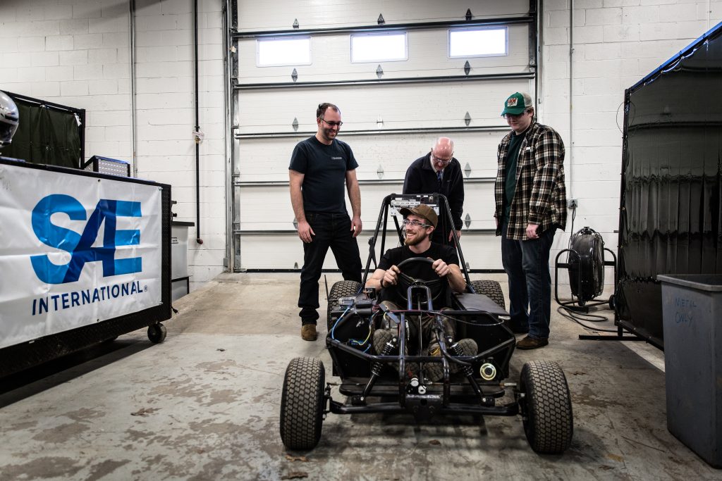 students standing around STEAM electric go kart. One student in driver seat smiling