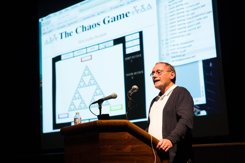 a speaker at a podium, speaking at a math-related event. A presentation slide titles The Choas Game is projected behind him